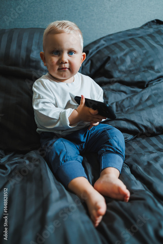 Obraz na płótnie Boy 2 years old with a smartphone. Toddler boy sitting in bed and playing with a mobile phone. Calling mom. Cute boy holding a mobile phone in his hands and looking intently at the screen. w salonie