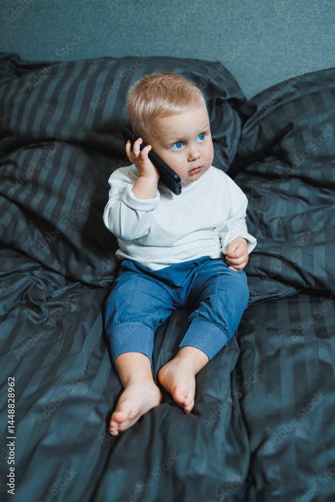 Cute little boy 2 years old using smartphone, looking at screen, child holding phone in hands, sitting on sofa at home alone, playing game on mobile device, watching cartoons online