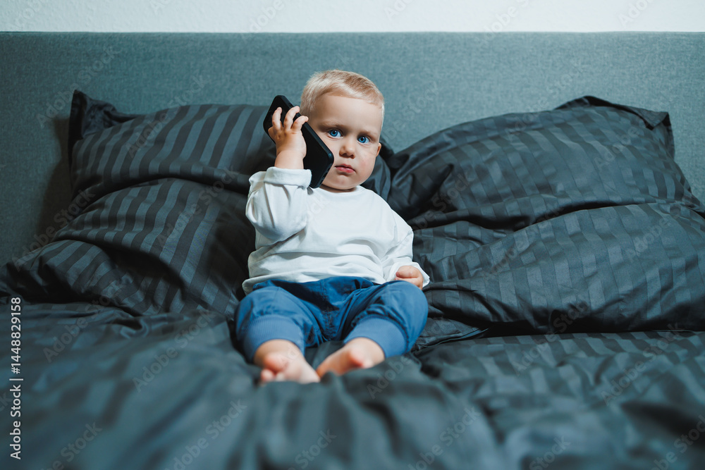 Boy 2 years old with a smartphone. Toddler boy sitting in bed and playing with a mobile phone. Calling mom. Cute boy holding a mobile phone in his hands and looking intently at the screen.
