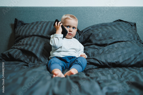 Obraz na płótnie Boy 2 years old with a smartphone. Toddler boy sitting in bed and playing with a mobile phone. Calling mom. Cute boy holding a mobile phone in his hands and looking intently at the screen. w salonie