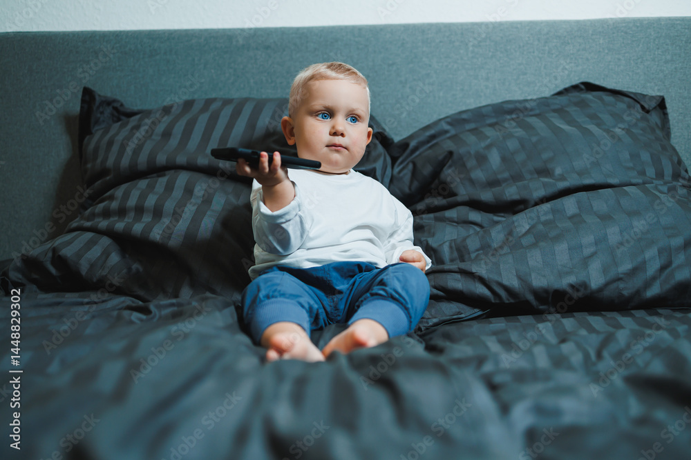 Toddler boy sitting on bed, deeply focused on playing on mobile phone. He is wearing a t-shirt and playing a game on his phone. Modern digitalization of children. Toddler with mobile phone