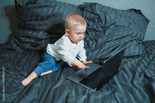 Cute boy 2 years old sits in bed and presses keys on laptop. Child sits and watches video on laptop. Modern development and digitalization of children.