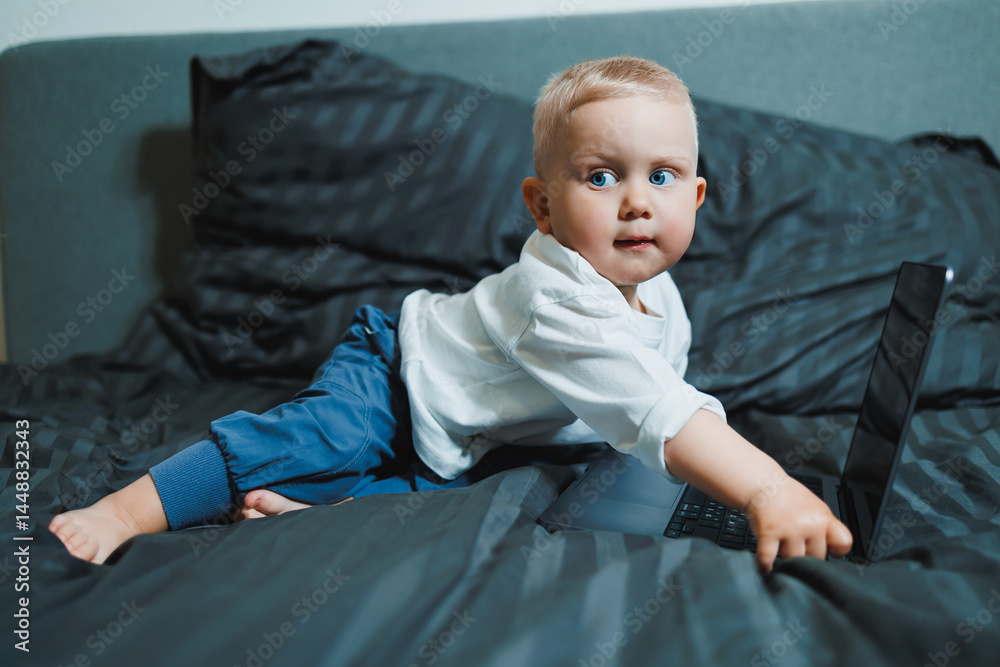 Little boy toddler using laptop watching video at home, early education and learning. Little child looking at laptop screen and listening to music while sitting on bed.
