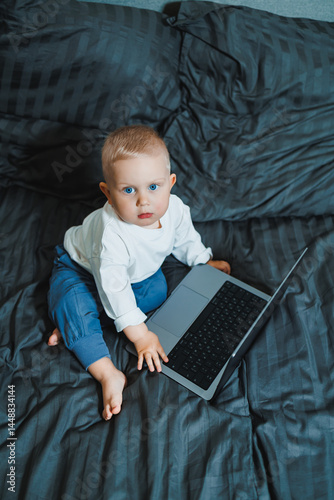 Obraz na płótnie Stylish blond toddler boy sitting on bed and playing with laptop at home in bedroom. Child with tablet computer watching cartoons. White cozy interior in bedroom. w salonie