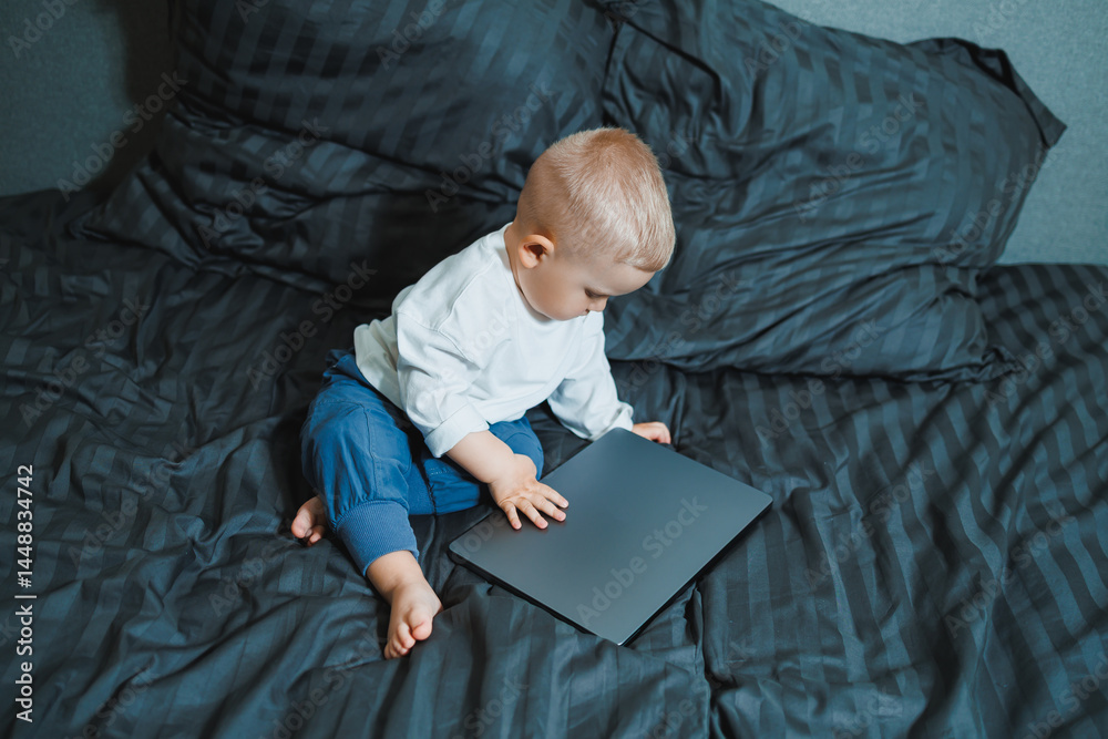 Stylish blond toddler boy sitting on bed and playing with laptop at home in bedroom. Child with tablet computer watching cartoons. White cozy interior in bedroom.