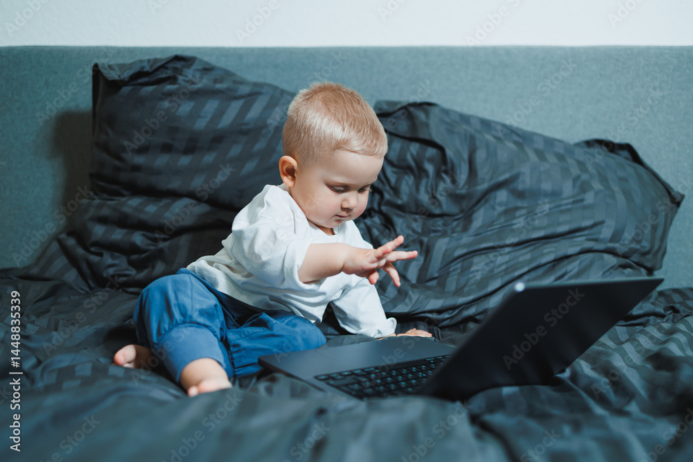 Stylish blond toddler boy sitting on bed and playing with laptop at home in bedroom. Child with tablet computer watching cartoons. White cozy interior in bedroom.