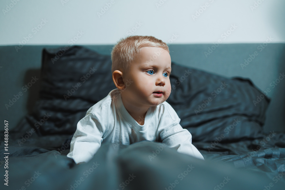 Child in bed. Cute toddler boy in white blouse and pants laughing while sitting in bed. 2 year old boy playing while sitting in bed. Home clothes for boys.