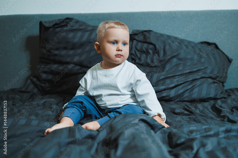 Two-year-old blue-eyed boy in a white long-sleeved shirt and pants sits in bed. Toddler boy in home clothes sits in the bedroom on the bed.