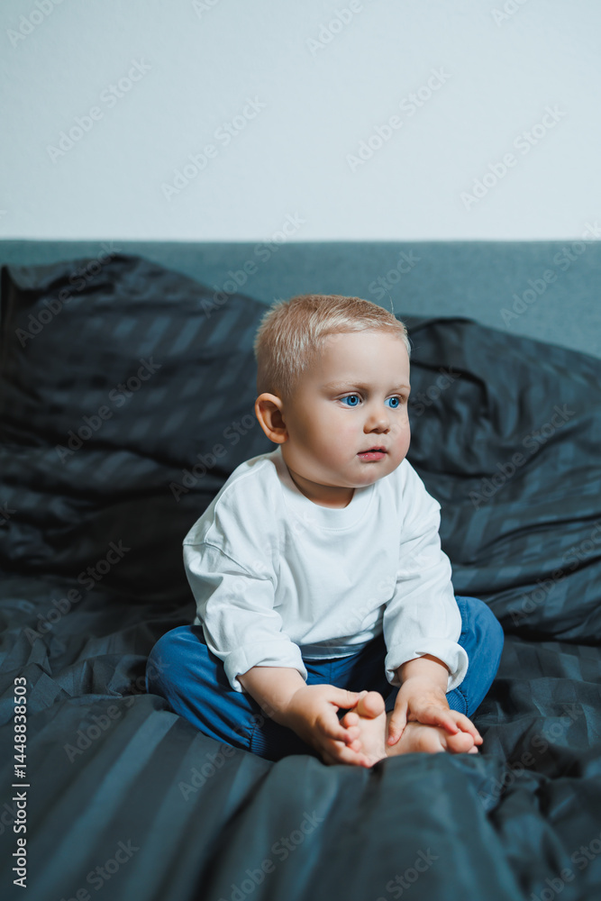 Child in bed. Cute toddler boy in white blouse and pants laughing while sitting in bed. 2 year old boy playing while sitting in bed. Home clothes for boys.