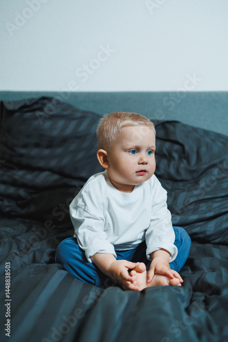 Child in bed. Cute toddler boy in white blouse and pants laughing while sitting in bed. 2 year old boy playing while sitting in bed. Home clothes for boys.