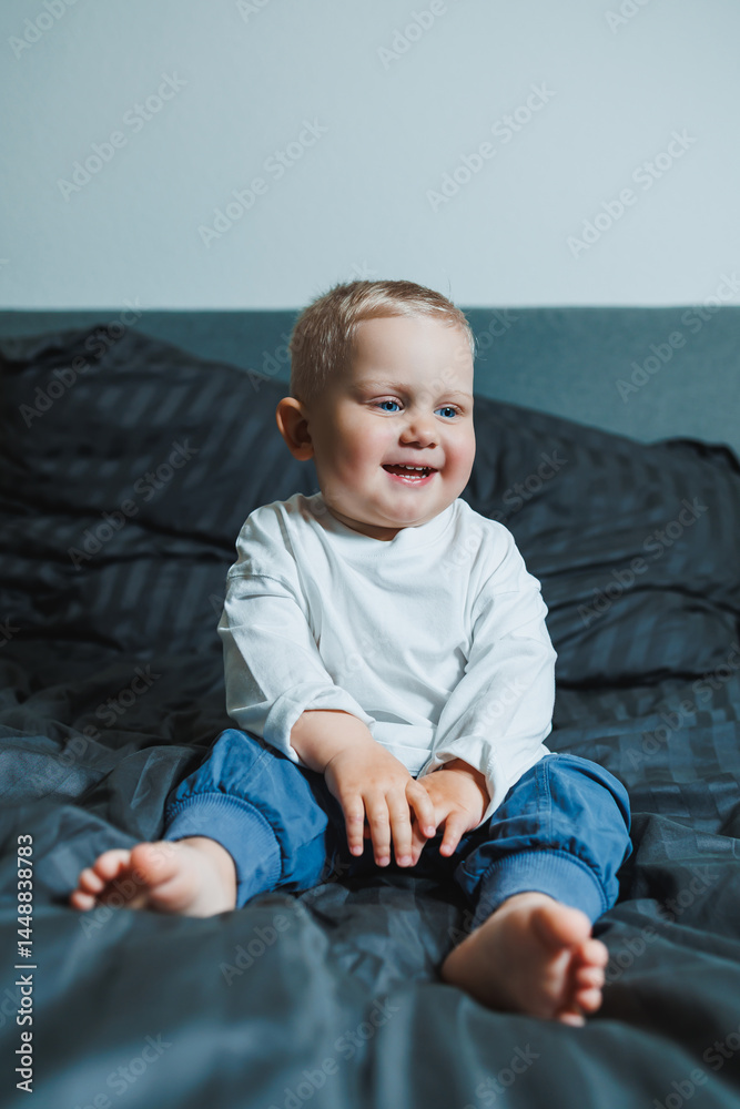 Cute toddler boy in white shirt and pants laughing while sitting in bed. 2 year old boy playing while sitting in bed. Home clothes for boys.