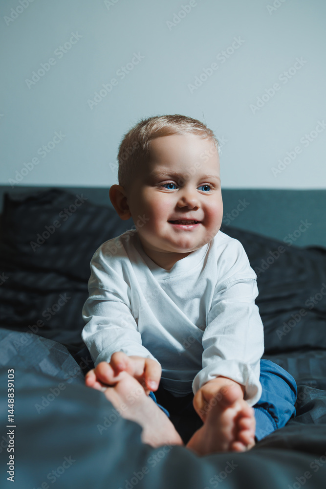 Cute toddler boy in white shirt and pants laughing while sitting in bed. 2 year old boy playing while sitting in bed. Home clothes for boys.