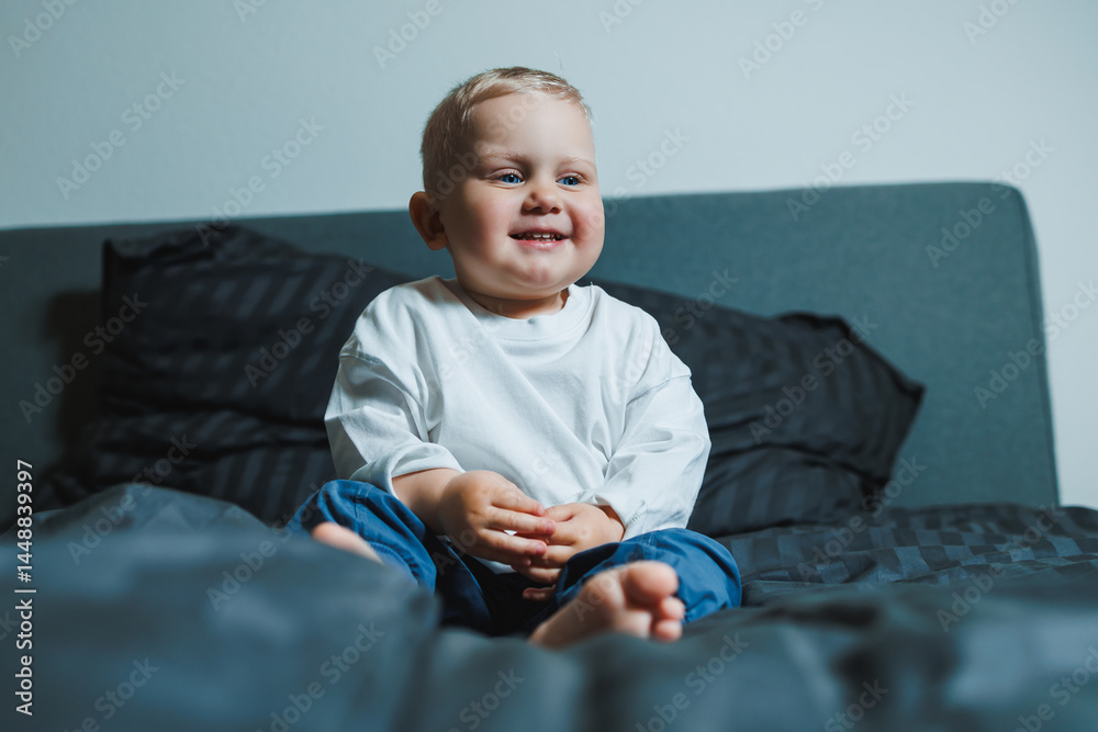 Cute toddler boy in white shirt and pants laughing while sitting in bed. 2 year old boy playing while sitting in bed. Home clothes for boys.