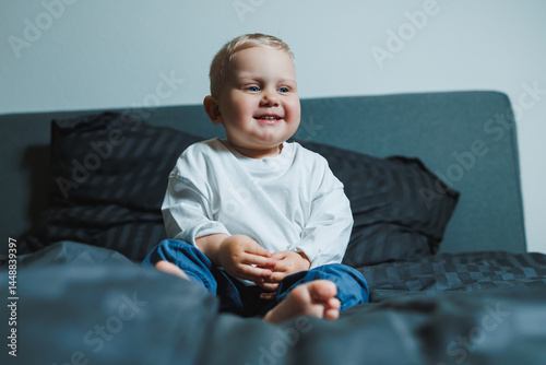 Obraz na płótnie Cute toddler boy in white shirt and pants laughing while sitting in bed. 2 year old boy playing while sitting in bed. Home clothes for boys. w salonie