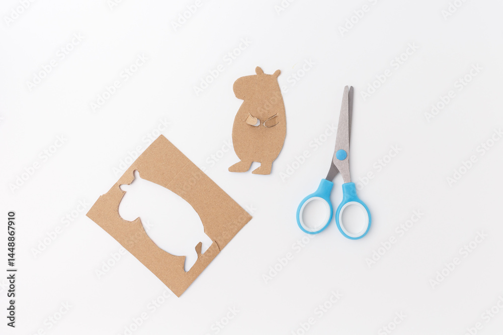 Fototapeta premium Cardboard capybara figure cut out with blue scissors on white background. DIY crafting process. Studio photo. Kids handmade art project and creative activity. part of project