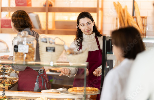 Obraz na płótnie Saleswoman in a red apron offers a customer fresh pastries with coffee, a coffee shop worker accepts an order for takeaway coffee and offers to take a delicious bun w salonie