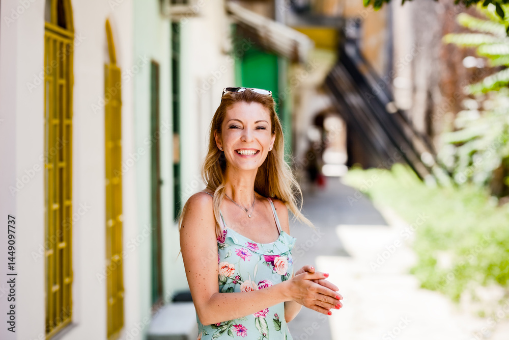 Cheerful Caucasian woman with blonde hair and a floral dress standing in a vibrant city alley, feeling happy and relaxed on a sunny summer day. Cheerful Caucasian woman with blonde hair and a floral dress standing in a vibrant city alley, feeling happy and relaxed on a sunny summer day.