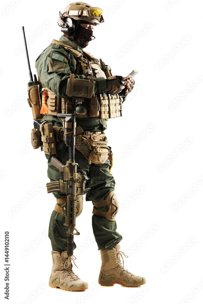 Fototapeta premium Armed soldier in full tactical gear holding a smartphone while standing against a white background