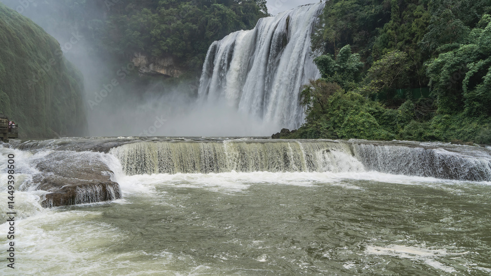 Fototapeta premium Beautiful tropical waterfall. Streams of white water flow down from the cliff. Foam, splashes, haze. Lush green vegetation. In the foreground is a riverbed with rapids. China. Huangguoshu Waterfall. 