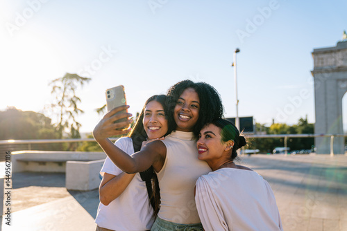 Three cheerful multi ethnic young women taking a selfie with smartphone in madrid city center with famous monument in background