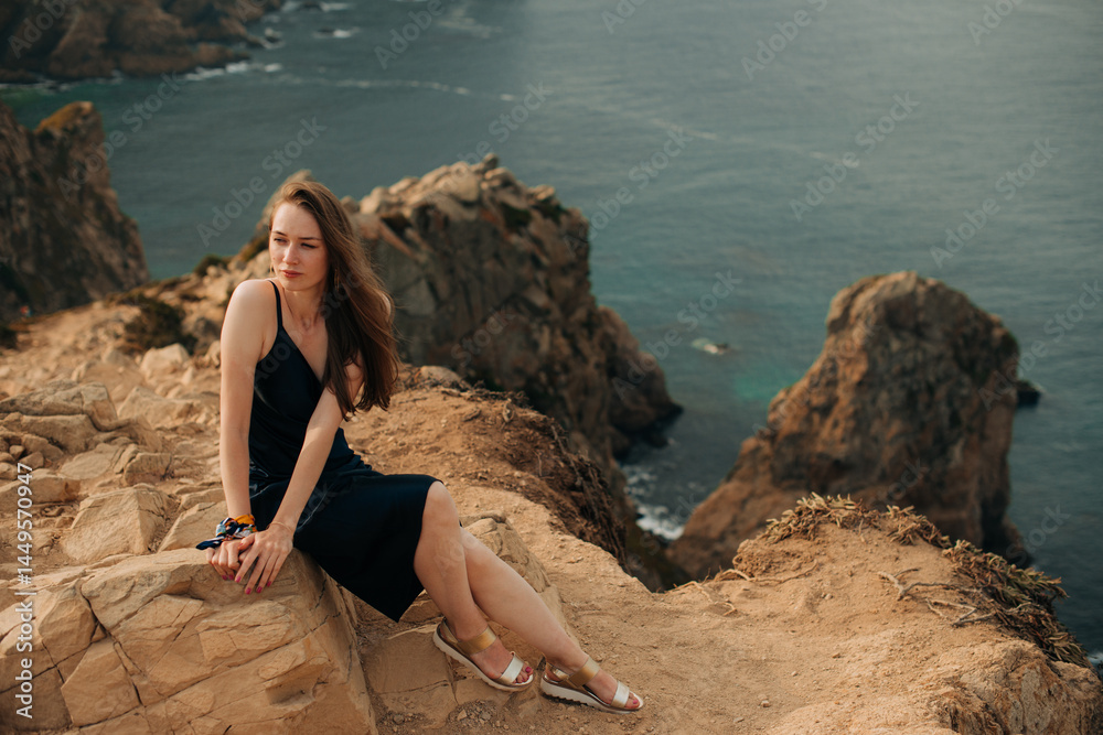 Woman in dark dress sitting on rocky cliff above ocean Woman in dark dress sitting on rocky cliff above ocean
