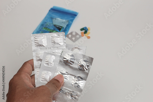 A hand holding several blister packs of medicine with scattered pills in the background, illustrating medication use, healthcare routine, or drug consumption concept.