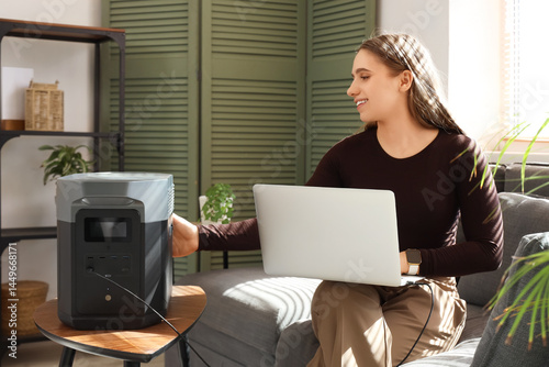 Young woman charging laptop with portable power station on sofa at home
