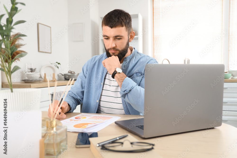 Young bearded man working with diagrams and laptop at table in kitchen Young bearded man working with diagrams and laptop at table in kitchen