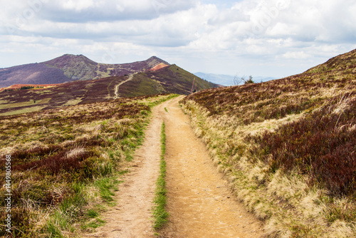 Fototapeta Naklejka Na Ścianę i Meble -  Scenic view of a winding dirt path through hills with lush greenery under a partly cloudy sky. Perfect for outdoor recreation and nature enthusiasts.