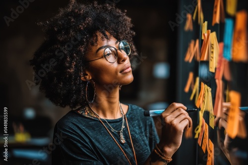 A focused young woman with glasses is brainstorming ideas on a glass board with colorful sticky notes, holding a marker and looking thoughtfully at the notes.