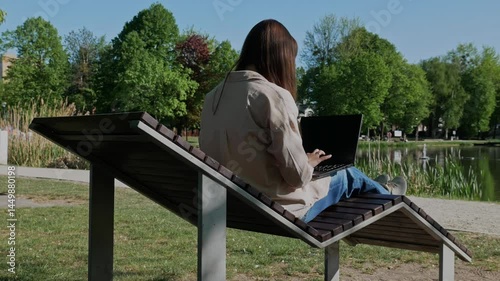  A modern businesswoman works on her laptop in the open air, sitting on a lounge chair by the lake. Work and relaxation in harmony.