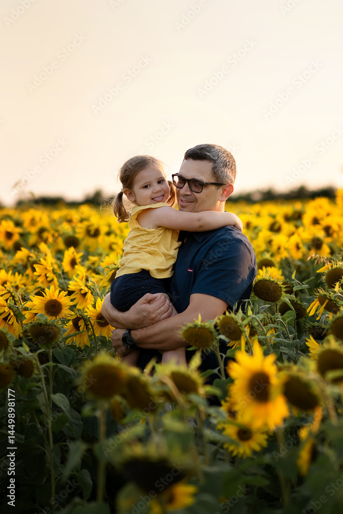 Fototapeta premium Happy laughing father holding baby daughter, standing together in sunflowers meadow. enjoying summer walking together on father's day.