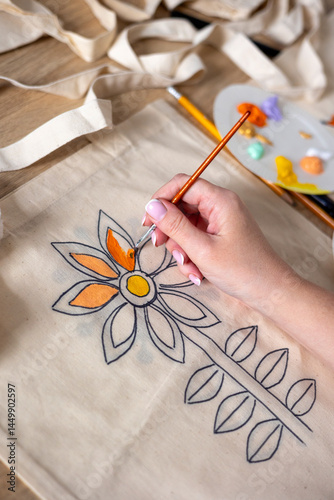 Woman's hand painting a flower on a canvas tote bag using acrylic paints. Close-up of a female hand painting a floral pattern on a reusable canvas bag with orange and yellow acrylic paints. Reuse
