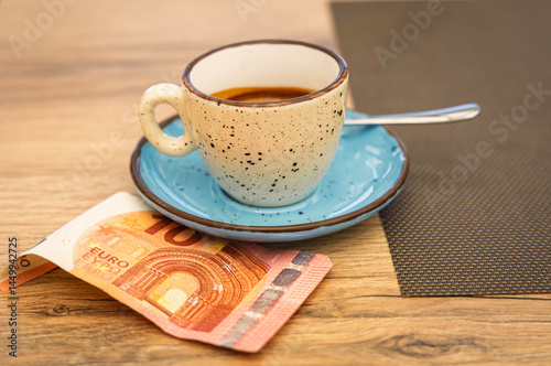A cup of espresso on a rustic saucer with a 10 euro banknote underneath, placed on a wooden table. Concept of tipping, hospitality, or café culture in Europe.