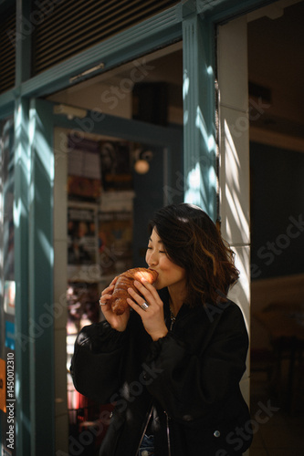 Asian woman enjoying a croissant at an outdoor cafe. Casual lifestyle moment, street food vibes, urban breakfast scene, tasty pastry and relaxing atmosphere.