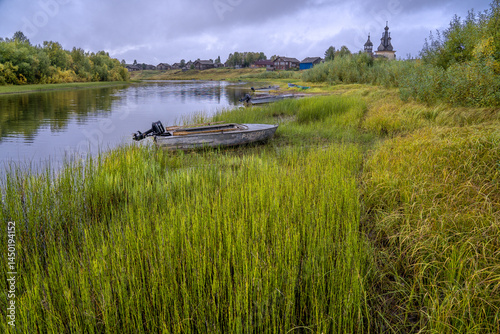 Boats on the river bank against the backdrop of log houses and an old wooden church.