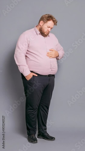 Overweight man with beard wearing a pink shirt and dark pants with hand on belly, expressing pain or discomfort.