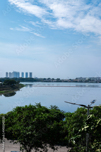 A calm lake reflects the sky and distant buildings, framed by green foliage. Serene and expansive.