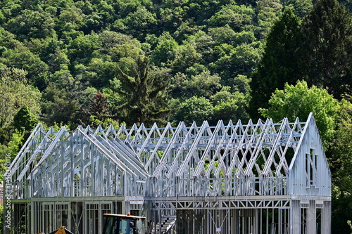 galvanized metal stud exterior wall frame for residential building. sloped roof. openings for windows. house construction process. lush green background. trees on a hill side. modern frame structure