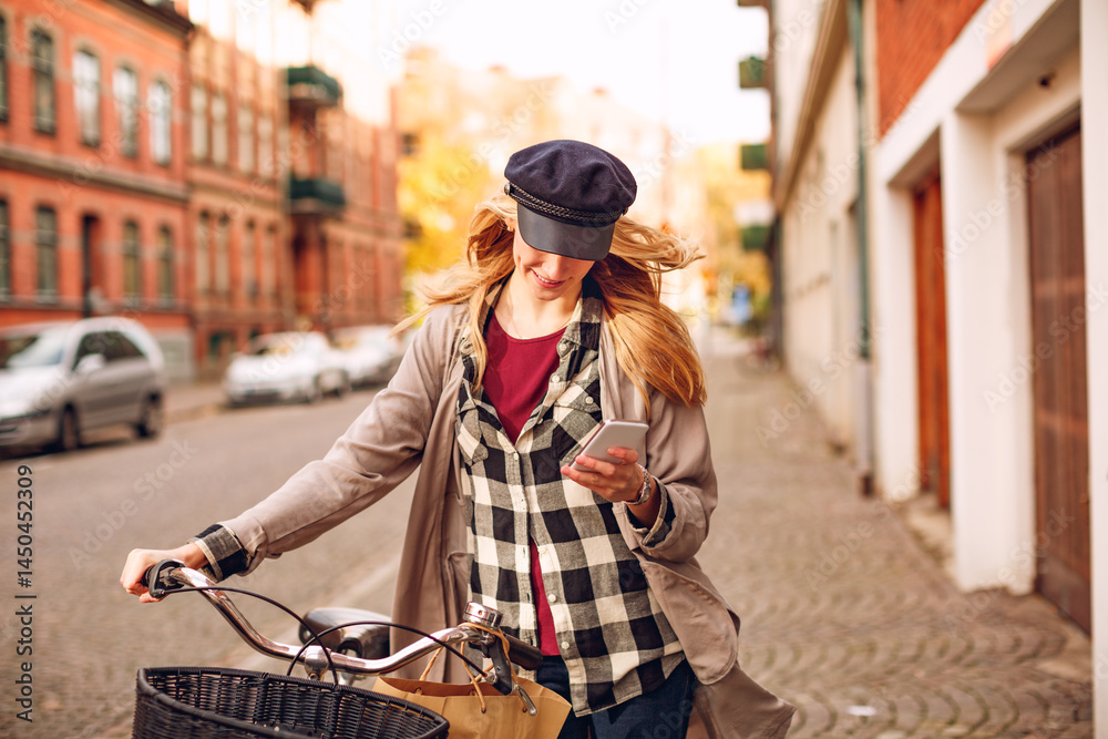 Fototapeta premium Young caucasian woman using a smart phone while pushing her bicycle on a city sidewalk