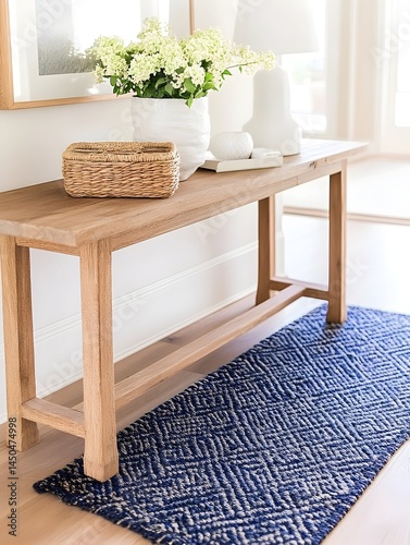 A long, light wood console table holds flowers, a basket, and books. A navy blue and beige geometric rug sits beneath