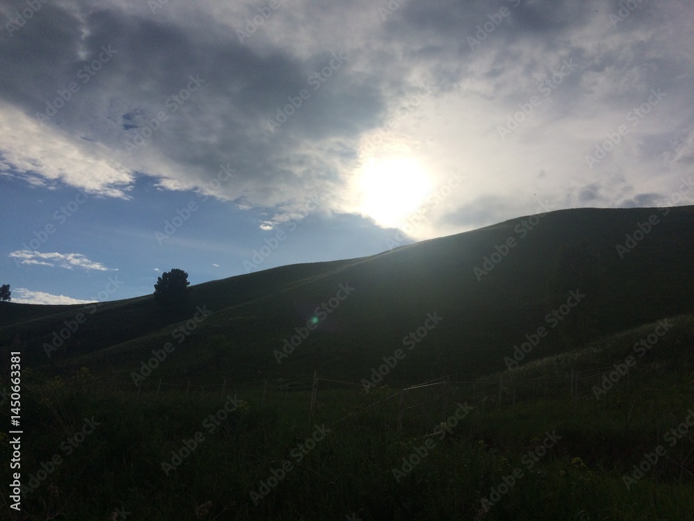 Fototapeta premium Sunlight peeks through clouds over rolling green hills at dusk near a serene landscape