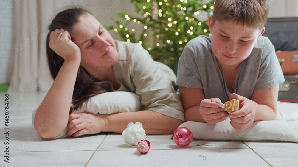 mom and son lying on marble floor close together surrounded by colorful christmas ornaments smiling warmly cozy festive home atmosphere decorated christmas tree in background bright joyful holiday