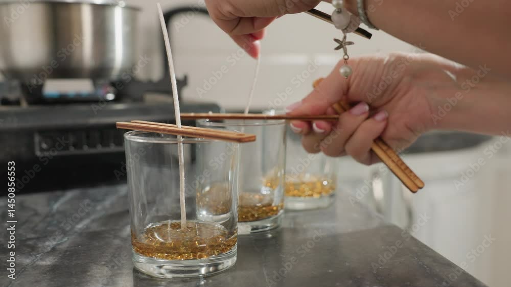 Close up of person using wooden chopsticks to secure candle wick firmly at center of glass cups filled with melted wax beads, with background view of pot on gas burner in modern kitchen setting Close up of person using wooden chopsticks to secure candle wick firmly at center of glass cups filled with melted wax beads, with background view of pot on gas burner in modern kitchen setting