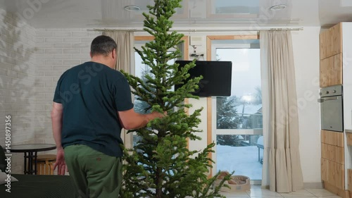 Wallpaper Mural Man in casual clothing rearranging and positioning artificial Christmas tree into perfect position near window, with snowy outdoor view, preparing for holiday season in cozy living room Torontodigital.ca