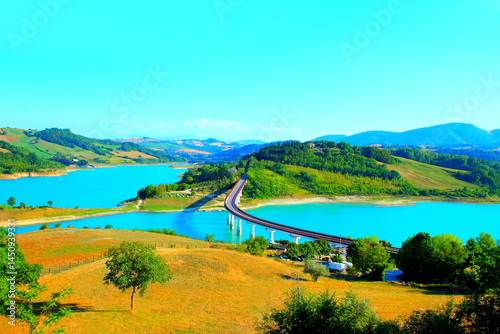 Picture-perfect scene from Lago di Cingoli, a complete view encompassing the whole breadth of the bridge, its deck and piers standing above the waters, surrounded by vegetation and the Sibillini