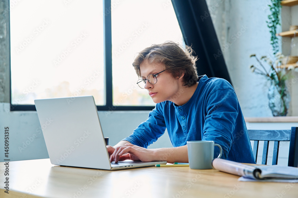 Teenage guy student sitting at the table with laptop, textbooks in home interior