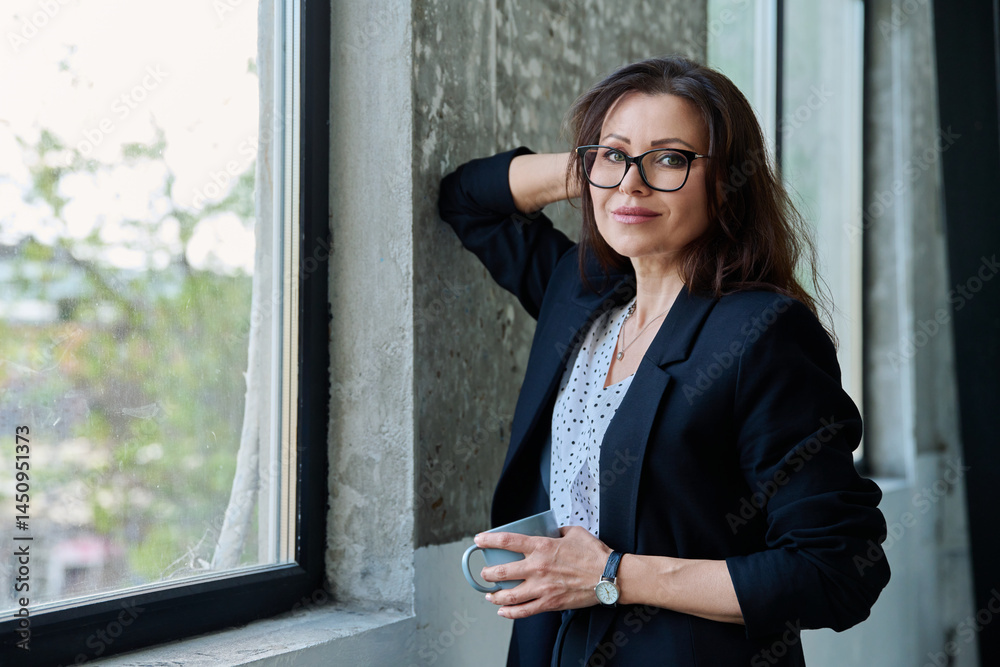 Portrait of middle aged beautiful businesswoman looking at camera, standing near window