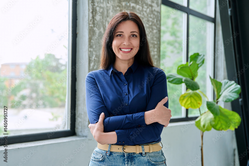 Portrait of beautiful confident brunette woman looking at camera with crossed arms, near window