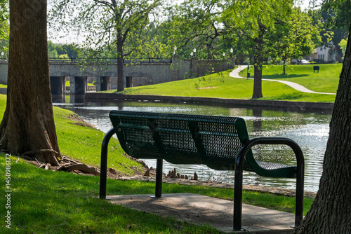 Bilde på lerret Empty park bench overlooking a calm pond and stone bridge